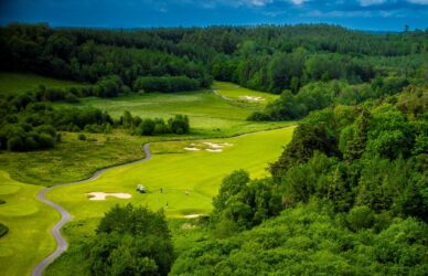 Scenic view of lush green golf course surrounded by dense forest under clear blue sky, perfect for leisure and relaxation.