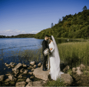 Couple embraces on lakeside rocks with lush green forest, clear blue skies; romantic wedding venue near serene waters.