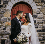 Romantic couple in elegant wedding attire share a moment by historic stone wall and rustic red door, enjoying scenic hotel backdrop.