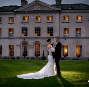 Elegant couple in wedding attire in front of lit-up manor hotel facade, lush lawn in foreground. Romantic wedding venue.